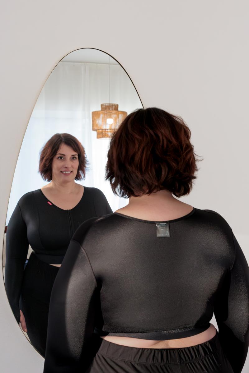 Frau in schwarzem Oberteil betrachtet sich im Spiegel, aufgenommen von hinten mit sichtbarem Spiegelbild.