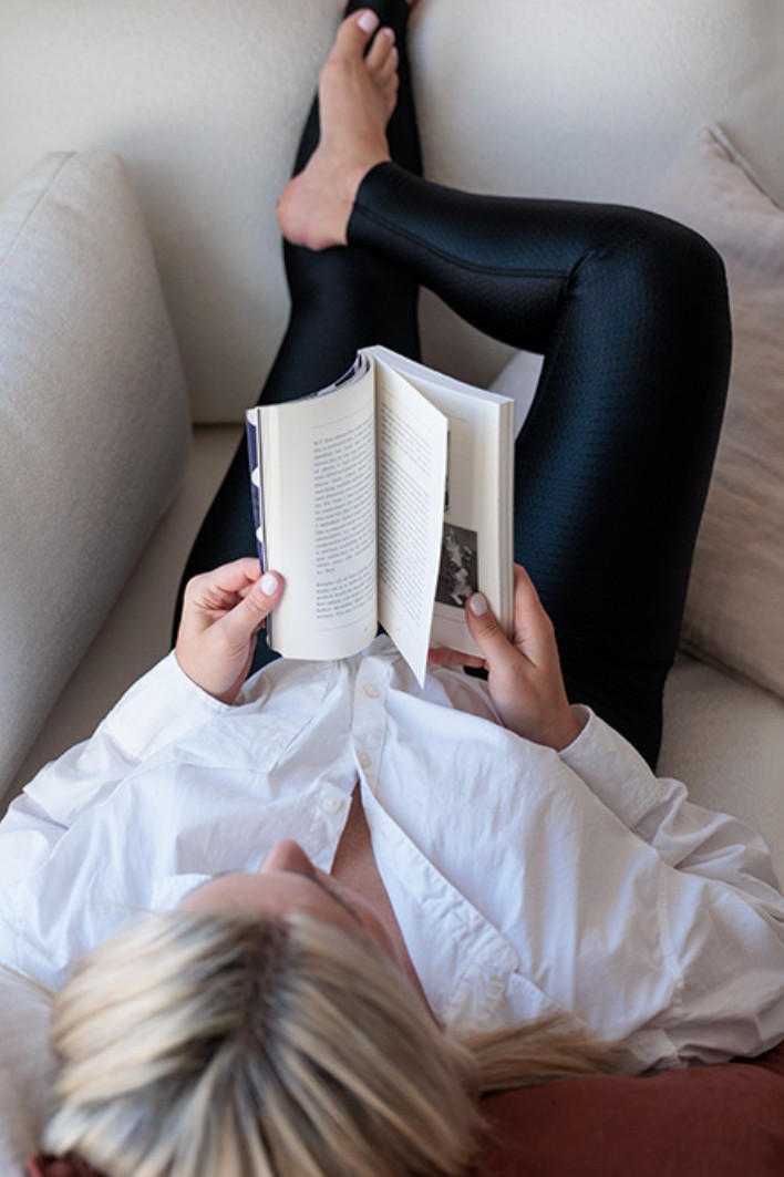 Frau liest ein Buch in der FLOW leggings auf dem Sofa, ihre Beine sind hochgelegt. 